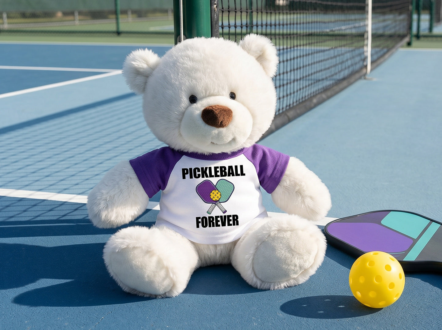 Personalized Pickleball Teddy Bear - 10" Creamy White Nuzzles Bear, Paddle Design - purple sleeved shirt, pictured on pickleball court. Perfect gift for pickleball enthusiasts.